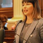 Woman in a gray blazer speaks into a microphone in a formal council chamber.