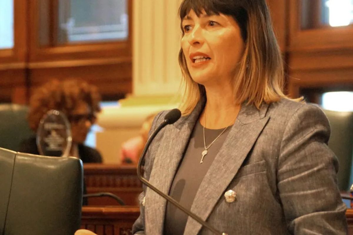 Woman in a gray blazer speaks into a microphone in a formal council chamber.