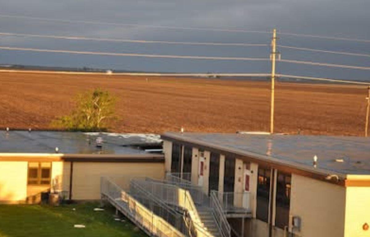 Sunlit exterior of a tan school building with a metal ramp and stairs along the front entrance under a cloudy sky.