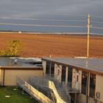 Sunlit exterior of a tan school building with a metal ramp and stairs along the front entrance under a cloudy sky.
