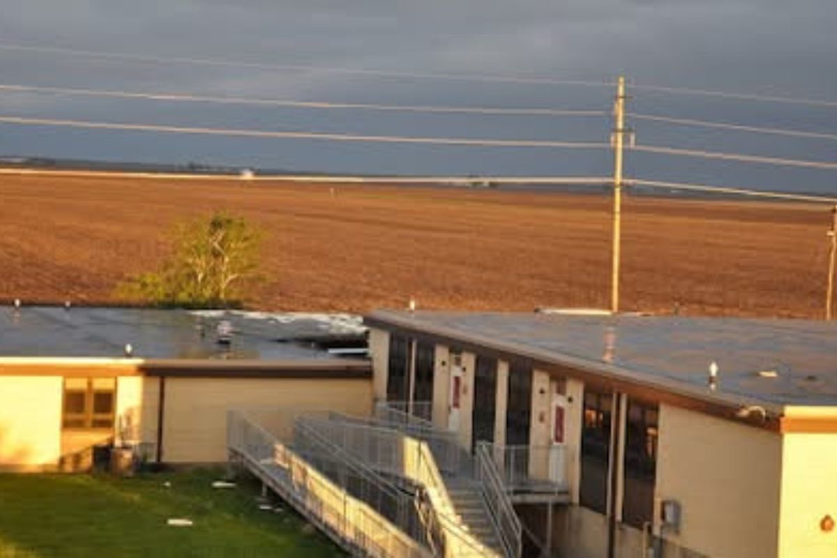 Sunlit exterior of a tan school building with a metal ramp and stairs along the front entrance under a cloudy sky.
