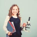 Smiling young girl student holds a microscope and several notebooks, ready for science class against a pale blue background.