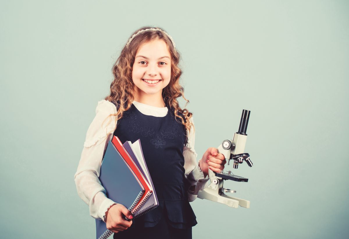 Smiling young girl student holds a microscope and several notebooks, ready for science class against a pale blue background.