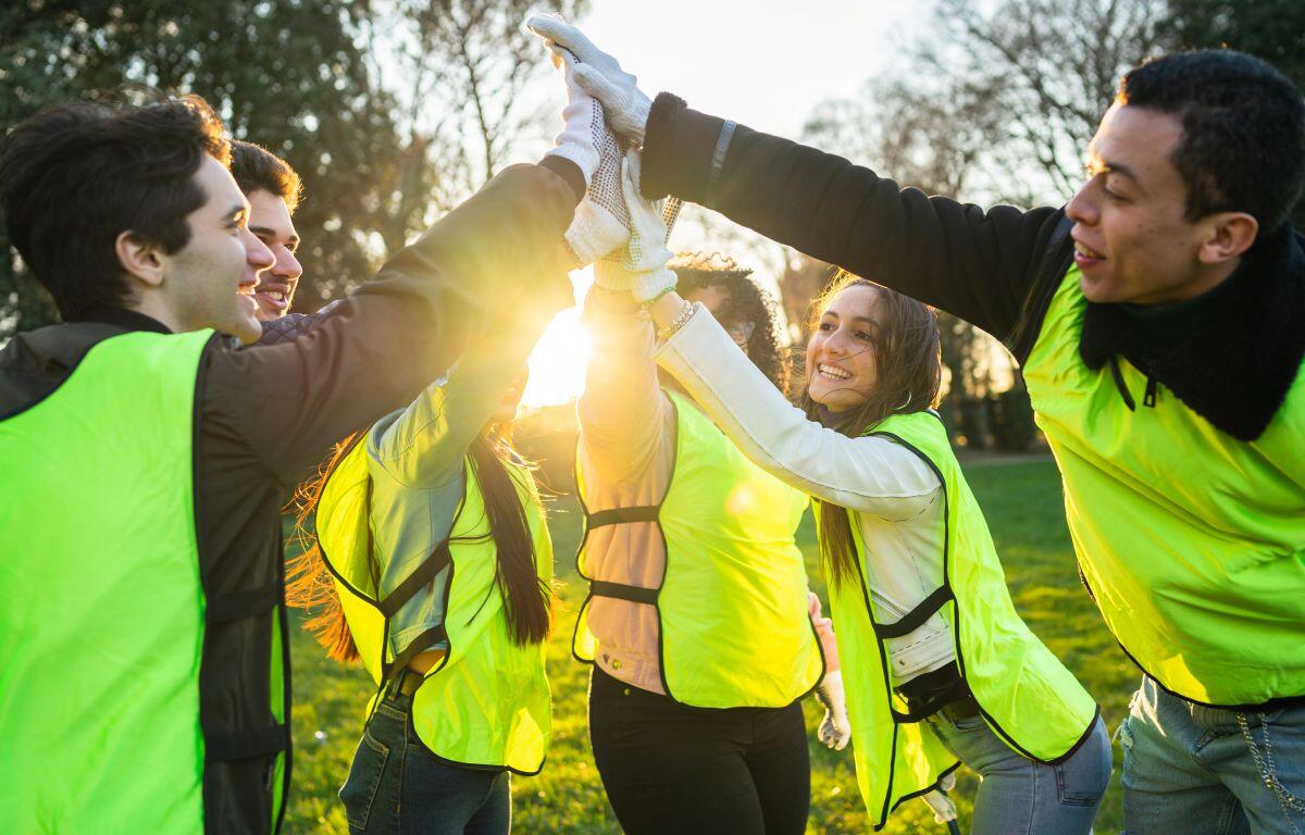 Group of diverse volunteers in neon vests cheerfully high-fiving outdoors at sunset in a park