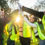 Group of diverse volunteers in neon vests cheerfully high-fiving outdoors at sunset in a park