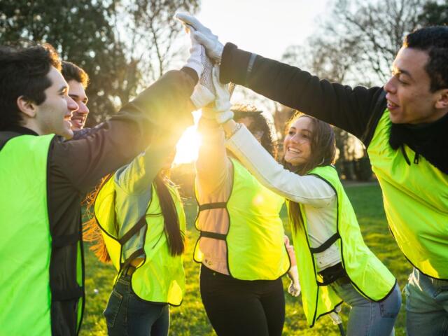 Group of diverse volunteers in neon vests cheerfully high-fiving outdoors at sunset in a park