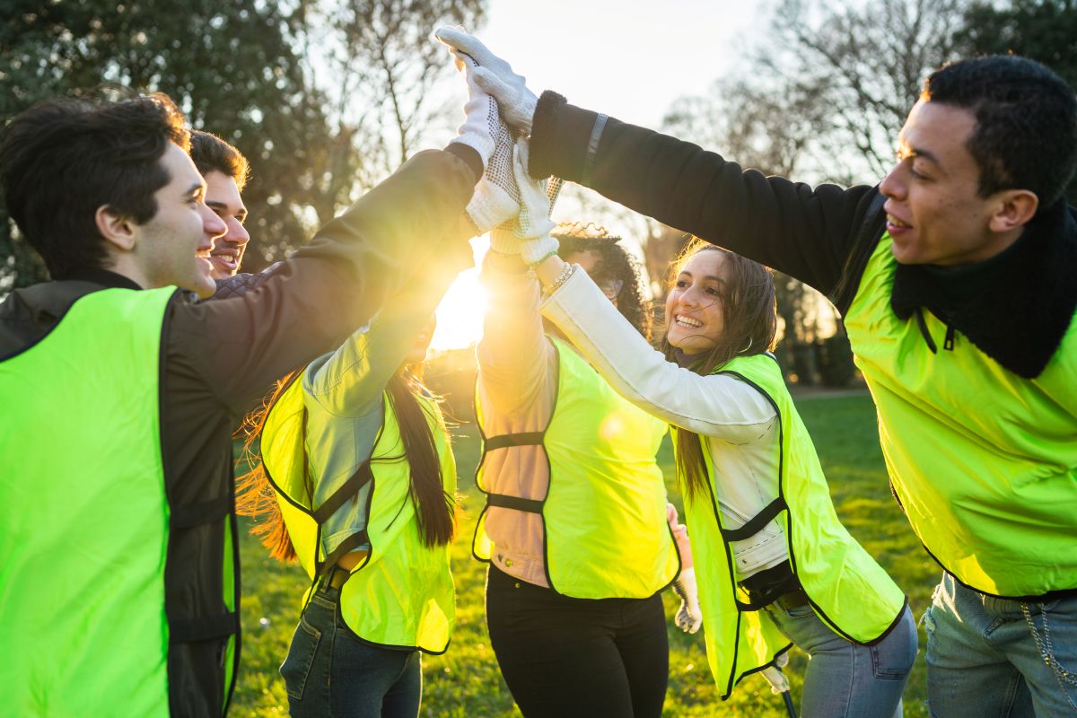 Group of diverse volunteers in neon vests cheerfully high-fiving outdoors at sunset in a park