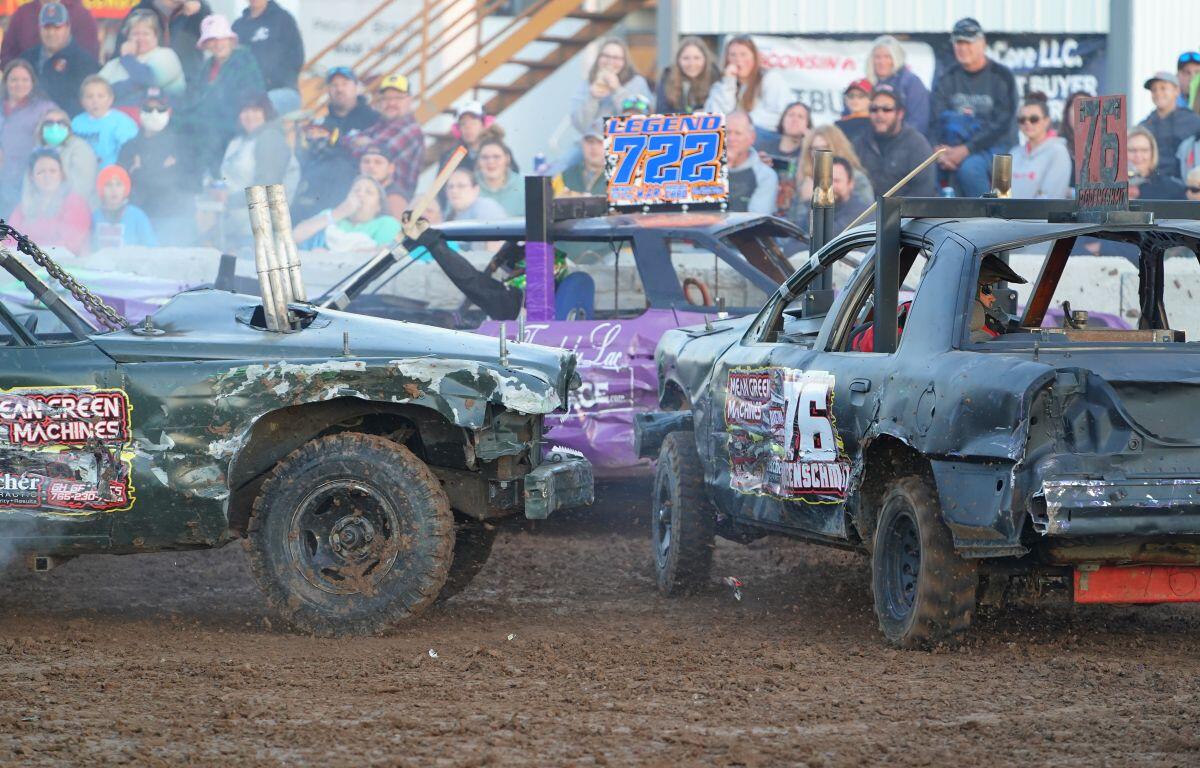 Two battered derby cars collide on a dirt track as a cheering crowd watches from the stands behind them, smoke rising slightly in the air.
