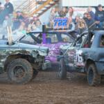 Two battered derby cars collide on a dirt track as a cheering crowd watches from the stands behind them, smoke rising slightly in the air.