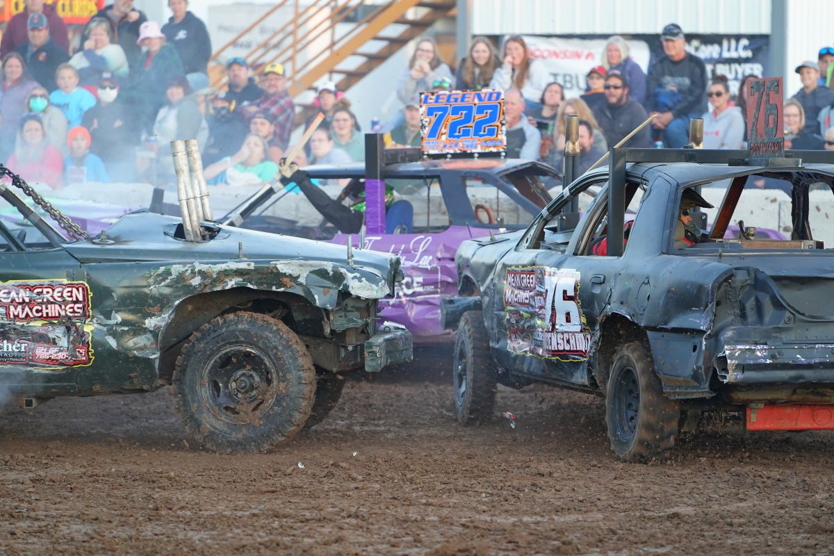 Two battered derby cars collide on a dirt track as a cheering crowd watches from the stands behind them, smoke rising slightly in the air.