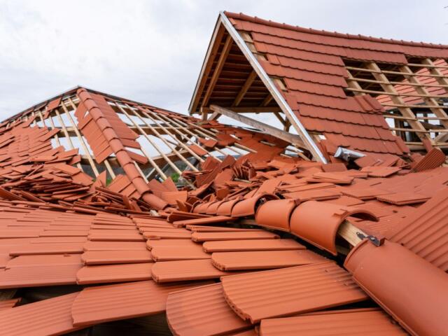 Collapsed red-tile roof with wooden framing exposed, debris scattered across the structure after damage.