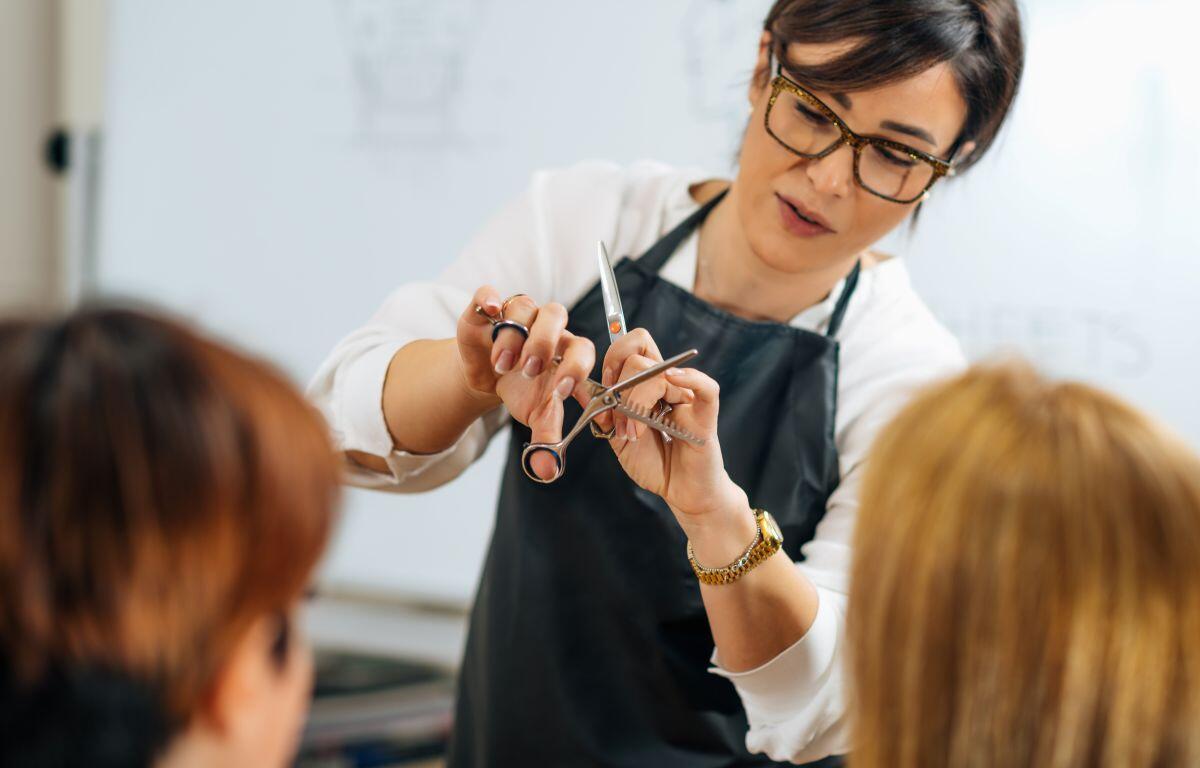 Hairdresser wearing glasses and a black apron cuts a client's hair with scissors and a comb in a salon.