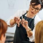 Hairdresser wearing glasses and a black apron cuts a client's hair with scissors and a comb in a salon.