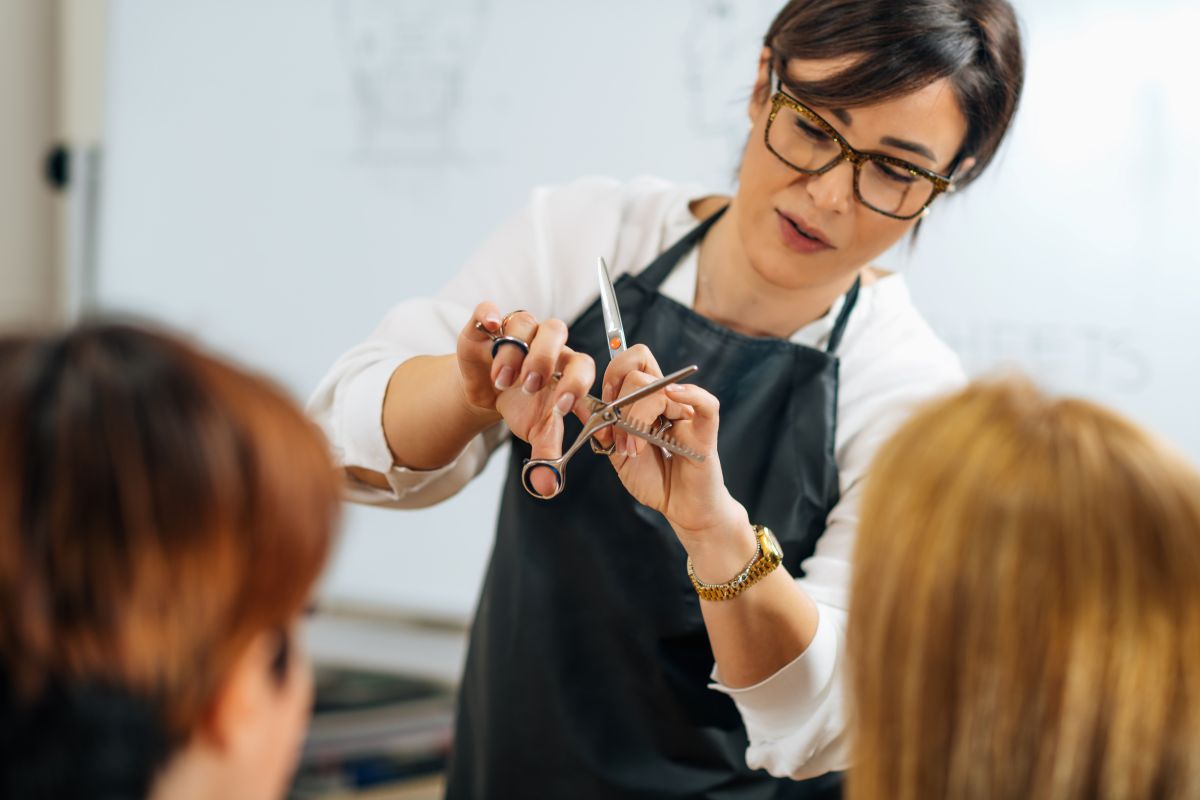 Hairdresser wearing glasses and a black apron cuts a client's hair with scissors and a comb in a salon.