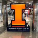 Large glowing orange numeral 1 on a dark base that says University of Illinois Urbana-Champaign in an airport terminal.