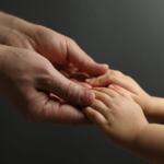 Adult hand gently cradles a baby's small hand, conveying care and connection against a dark background.