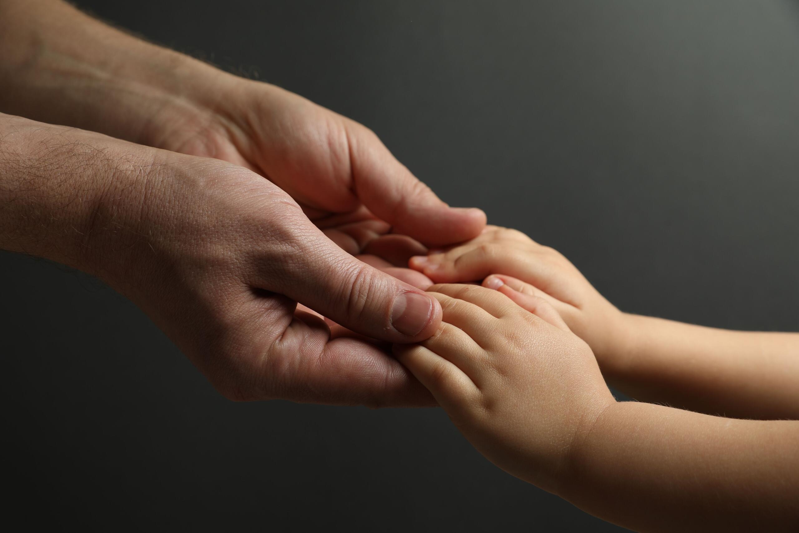 Adult hand gently cradles a baby's small hand, conveying care and connection against a dark background.