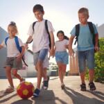 Group of four children with backpacks playing soccer on a sunny sidewalk, kicking a red-yellow ball together.