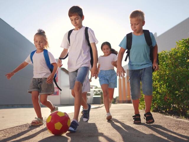 Group of four children with backpacks playing soccer on a sunny sidewalk, kicking a red-yellow ball together.