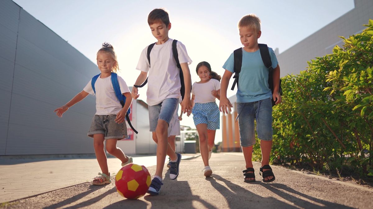 Group of four children with backpacks playing soccer on a sunny sidewalk, kicking a red-yellow ball together.