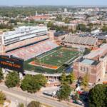Aerial view of a large college football stadium with orange and blue seating and a football field labeled Illinois Fighting Illini on the scoreboard.