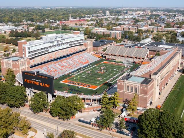 Aerial view of a large college football stadium with orange and blue seating and a football field labeled Illinois Fighting Illini on the scoreboard.