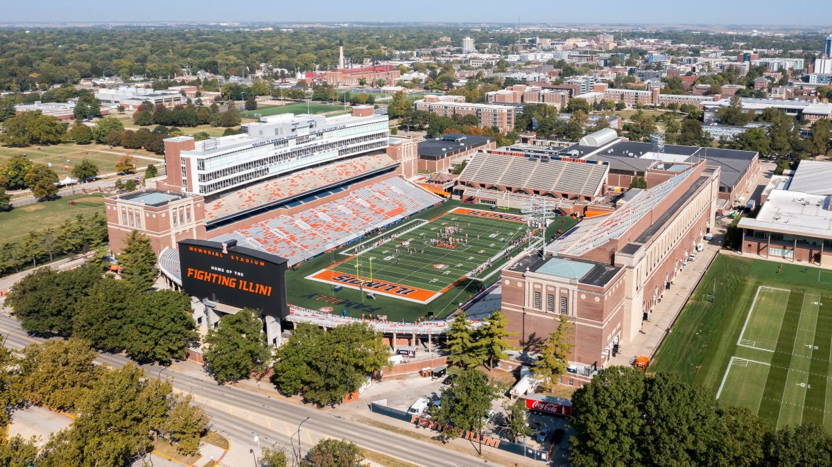 Aerial view of a large college football stadium with orange and blue seating and a football field labeled Illinois Fighting Illini on the scoreboard.