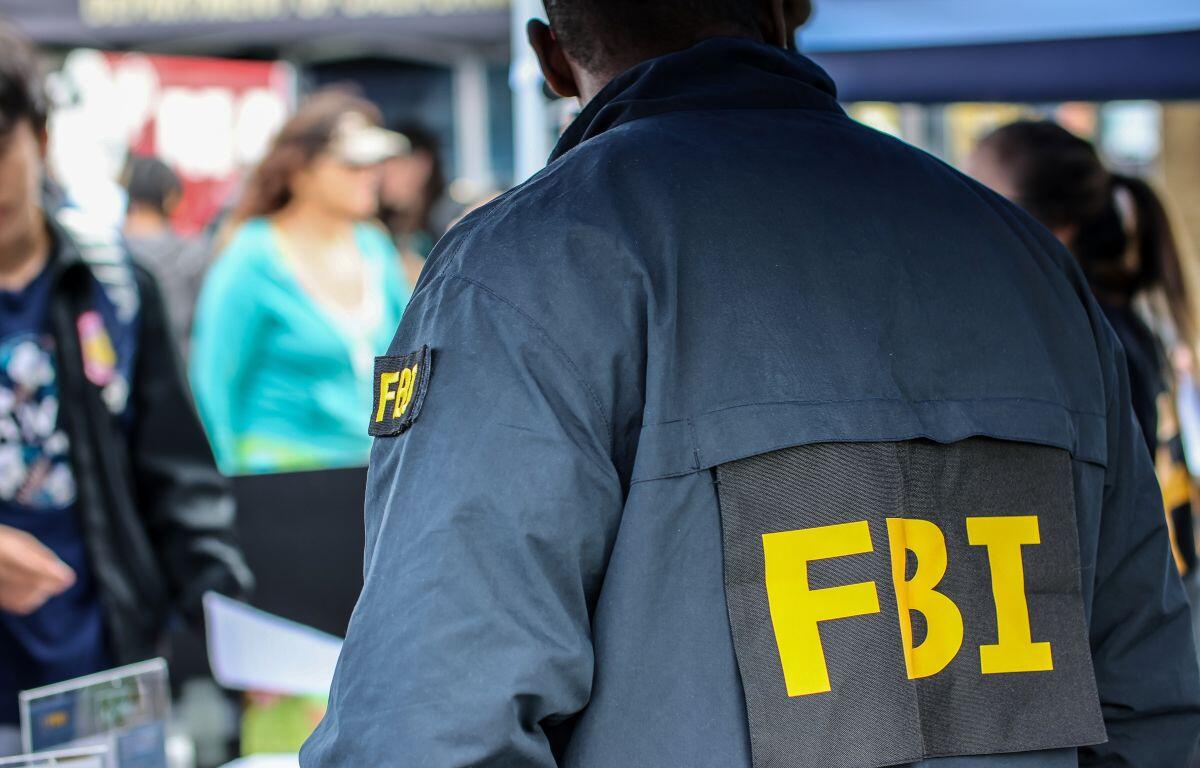 Back view of a person wearing a dark jacket with large yellow 'FBI' letters at an outdoor event, crowd in the background.