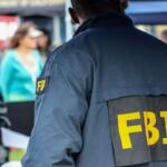 Back view of a person wearing a dark jacket with large yellow 'FBI' letters at an outdoor event, crowd in the background.