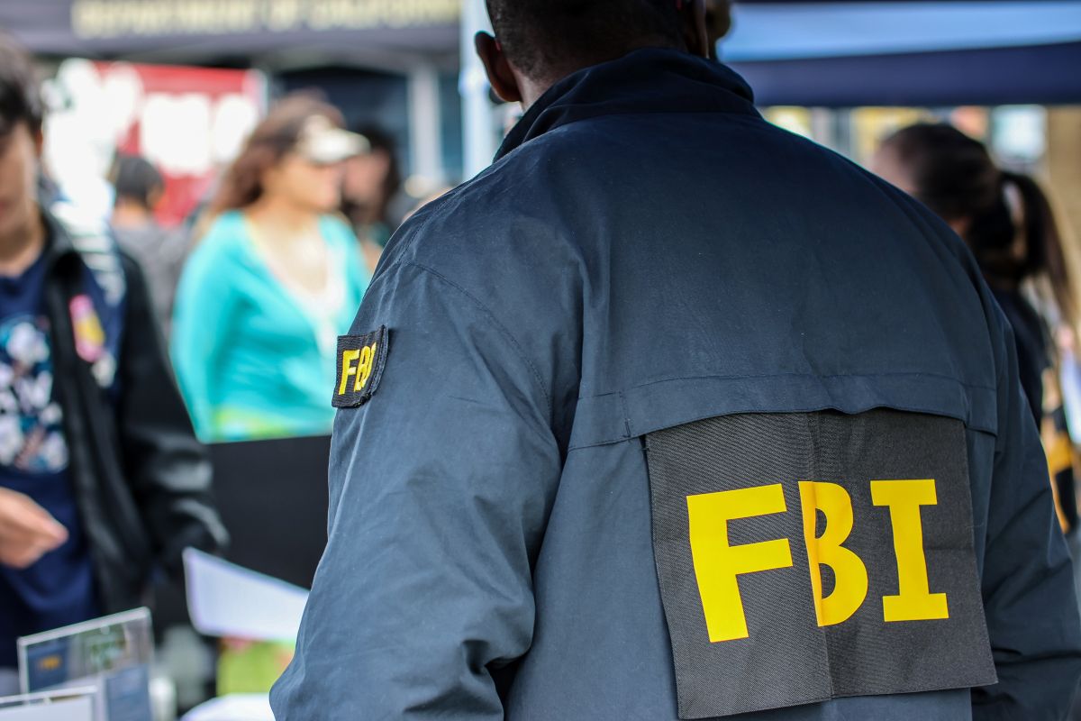 Back view of a person wearing a dark jacket with large yellow 'FBI' letters at an outdoor event, crowd in the background.