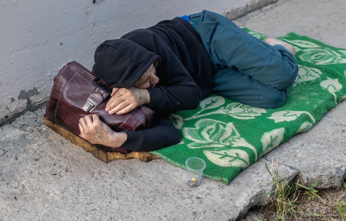 Person sleeping on a sidewalk under a hoodie, clutching a brown bag on a green patterned blanket.