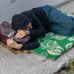Person sleeping on a sidewalk under a hoodie, clutching a brown bag on a green patterned blanket.