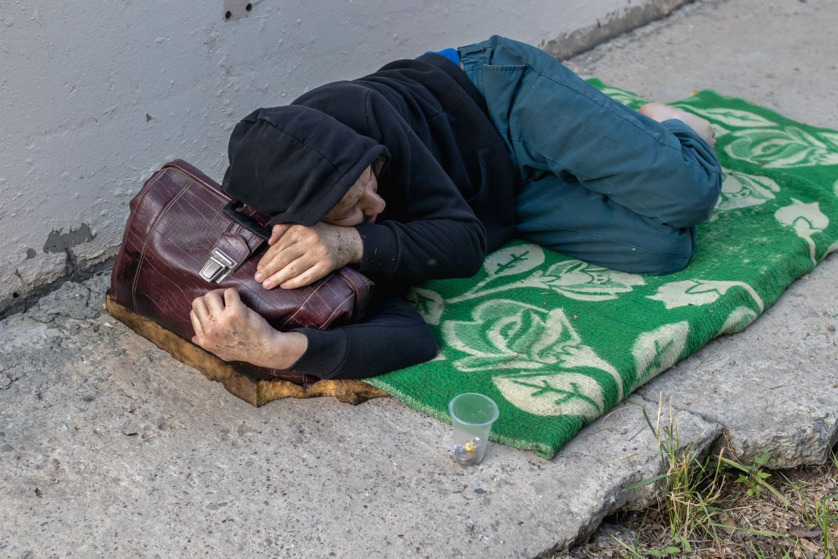 Person sleeping on a sidewalk under a hoodie, clutching a brown bag on a green patterned blanket.