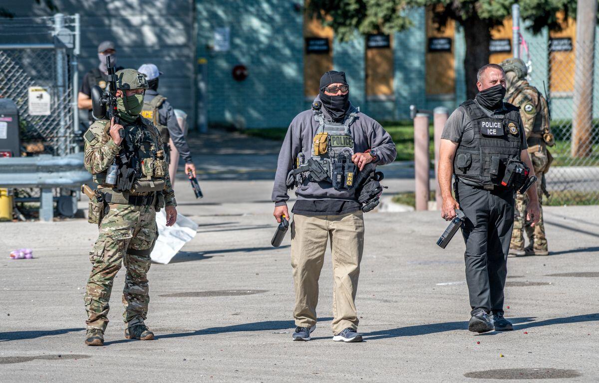 Tactical police team in armor and masks stand outdoors with weapons ready.