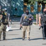 Tactical police team in armor and masks stand outdoors with weapons ready.