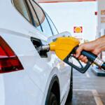 Person refueling a white car at a gas station with a yellow fuel nozzle in use, visible pump in background.