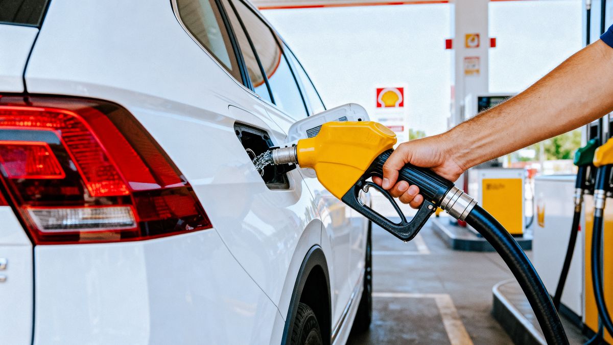 Person refueling a white car at a gas station with a yellow fuel nozzle in use, visible pump in background.