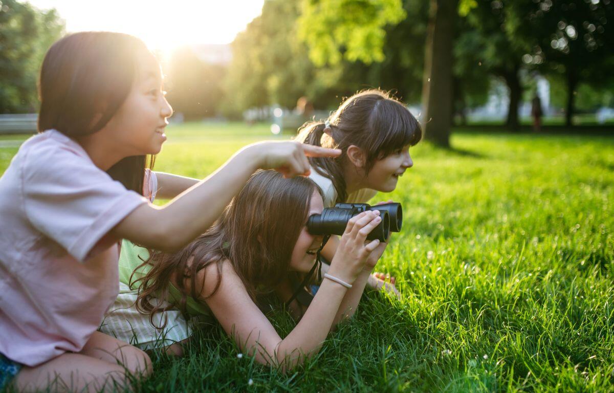 Three girls lying on green grass in a sunny park; one looks through binoculars while another points toward something in the distance, and a third watches nearby.