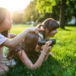 Three girls lying on green grass in a sunny park; one looks through binoculars while another points toward something in the distance, and a third watches nearby.
