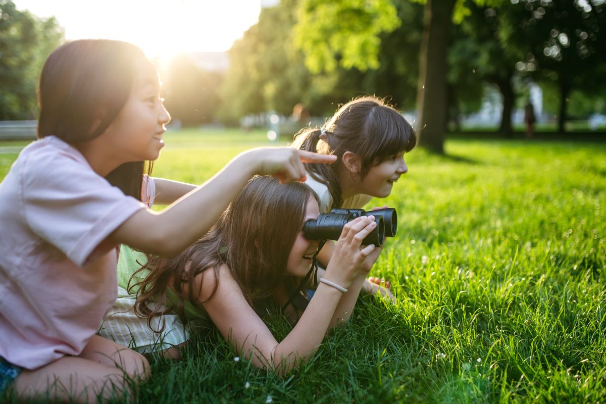 Three girls lying on green grass in a sunny park; one looks through binoculars while another points toward something in the distance, and a third watches nearby.