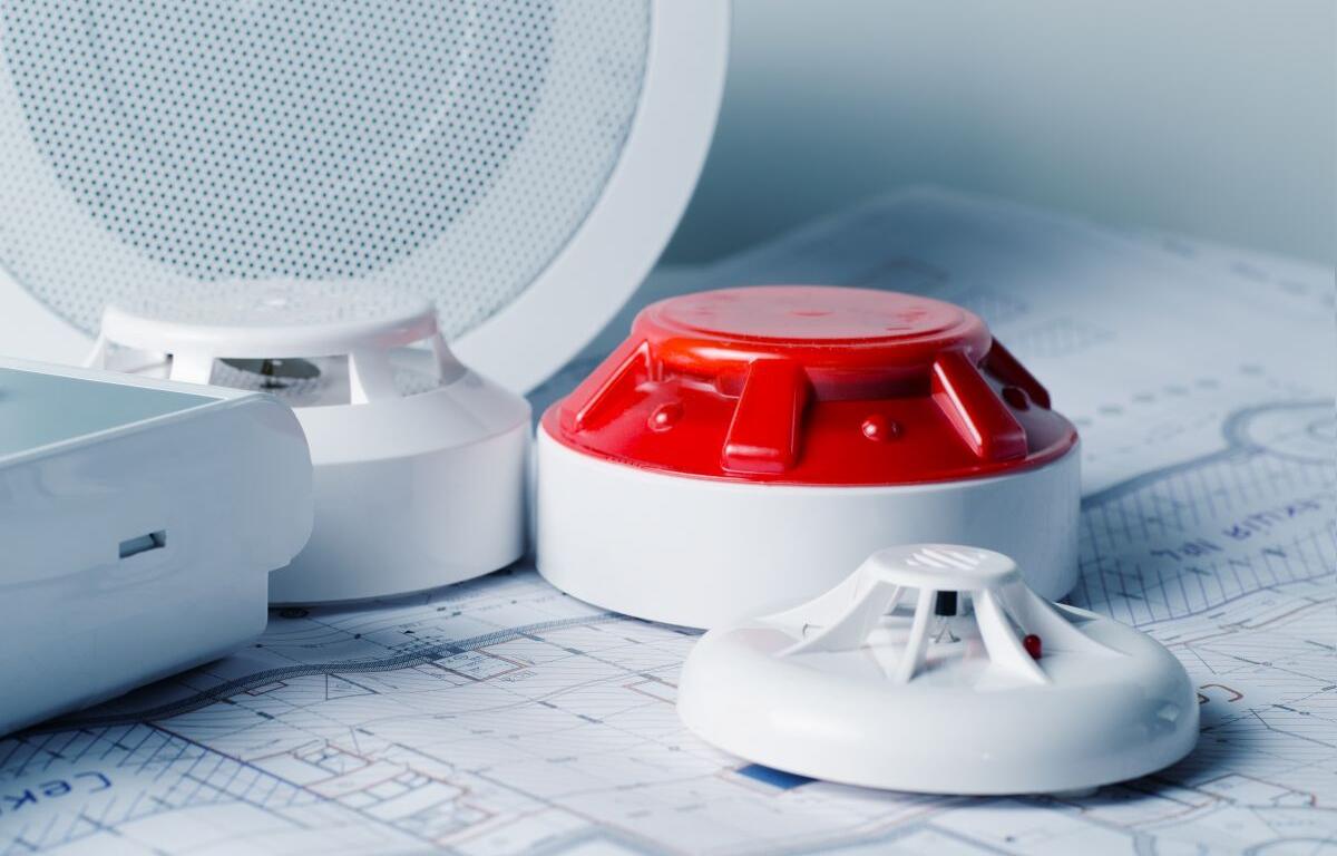 Smoke alarm components (red alarm unit, white base) and a smoke detector resting on architectural blueprints, suggesting fire safety planning.