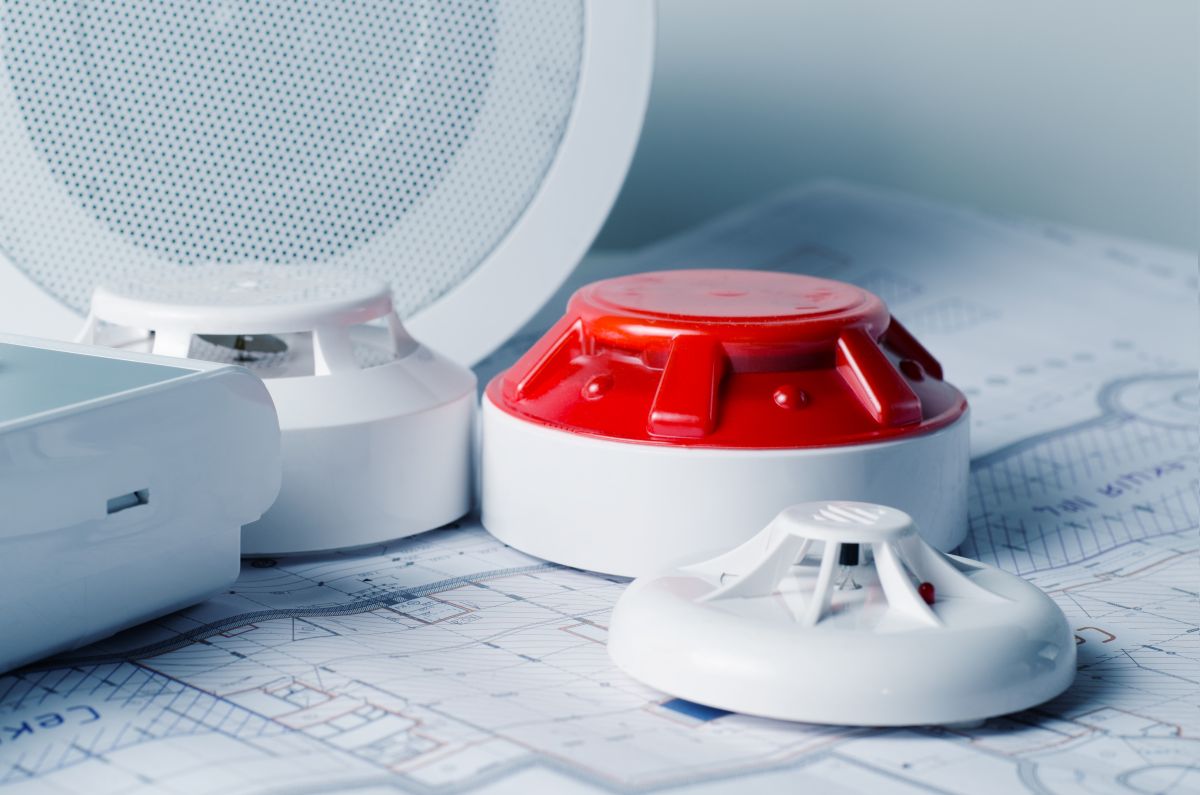 Smoke alarm components (red alarm unit, white base) and a smoke detector resting on architectural blueprints, suggesting fire safety planning.
