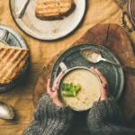 A person in a gray sweater holding a bowl of creamy soup garnished with parsley at a rustic table, with bread nearby.