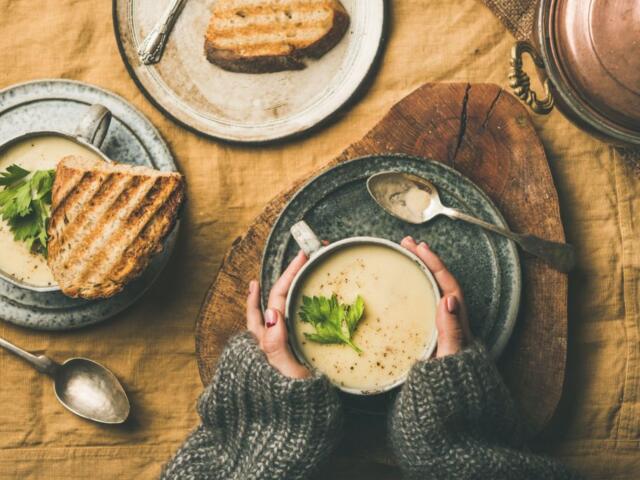 A person in a gray sweater holding a bowl of creamy soup garnished with parsley at a rustic table, with bread nearby.