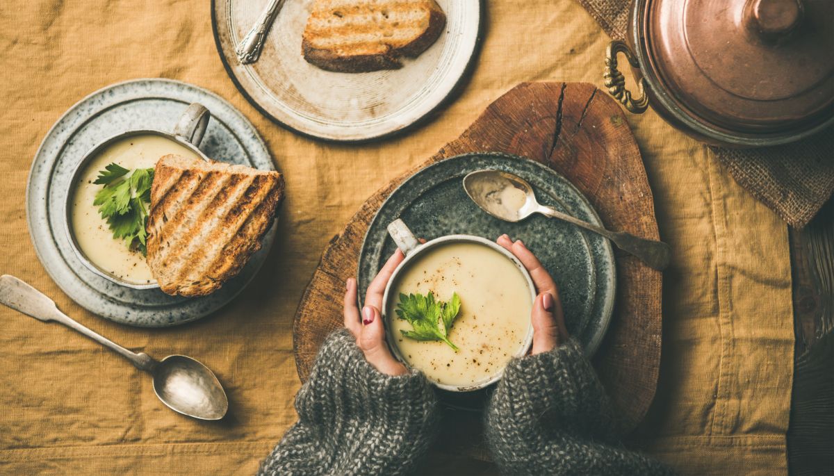 A person in a gray sweater holding a bowl of creamy soup garnished with parsley at a rustic table, with bread nearby.
