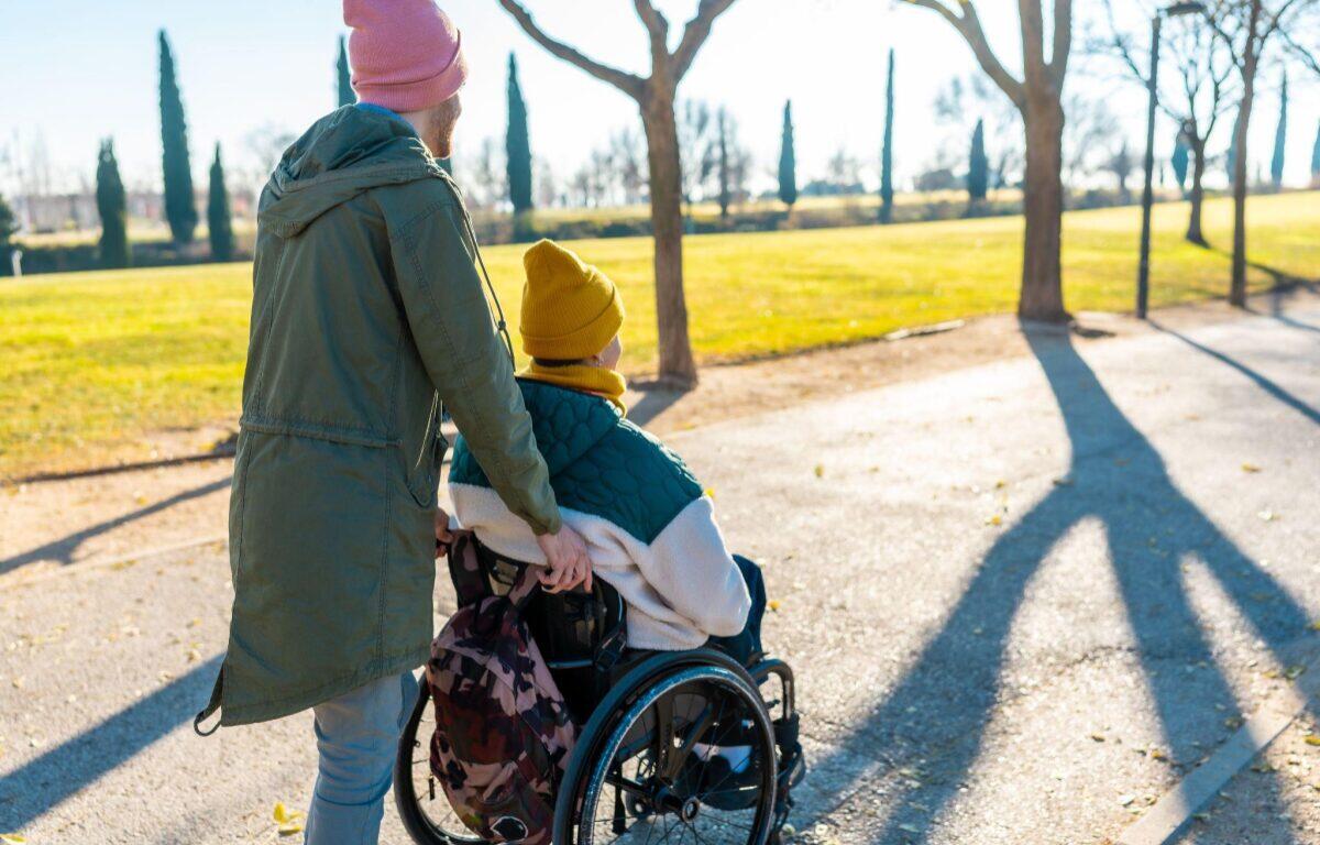 Adult pushing a person in a wheelchair along a park path on a sunny day; both wear knit hats and jackets, backpack attached to chair.