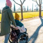 Adult pushing a person in a wheelchair along a park path on a sunny day; both wear knit hats and jackets, backpack attached to chair.