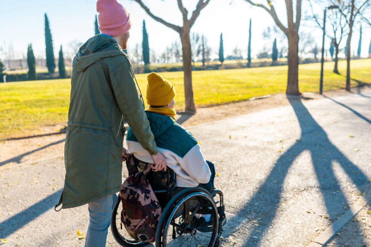 Adult pushing a person in a wheelchair along a park path on a sunny day; both wear knit hats and jackets, backpack attached to chair.