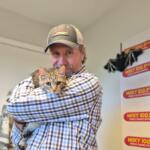 Man in a plaid shirt and cap hugs a tabby cat inside a radio studio with WIXY 100.3FM banners in the background.