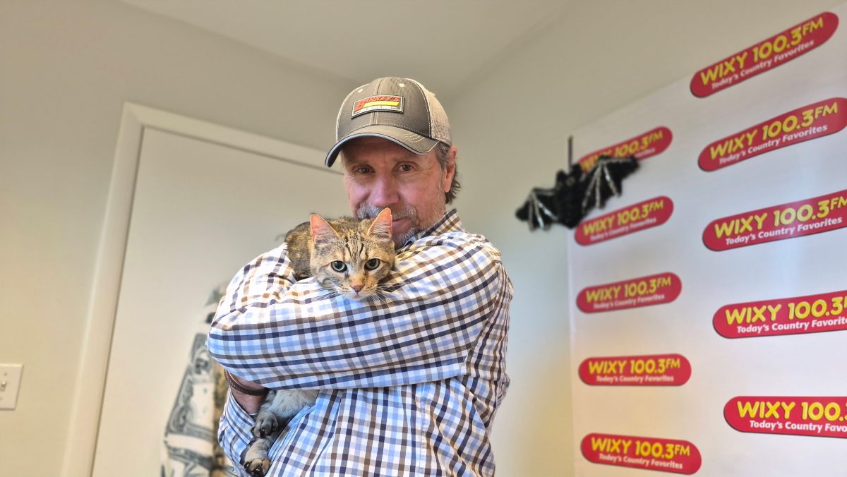Man in a plaid shirt and cap hugs a tabby cat inside a radio studio with WIXY 100.3FM banners in the background.